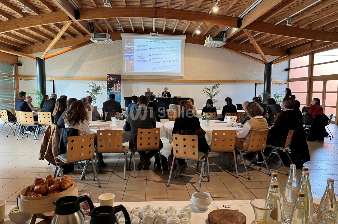 Participants assis dans une salle de conférence lumineuse, écoutant une présentation projetée sur un écran.