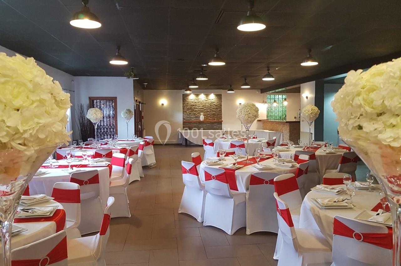 Salle de réception décorée avec des tables dressées, nappes blanches, chaises ornées de rubans rouges et centres de table…