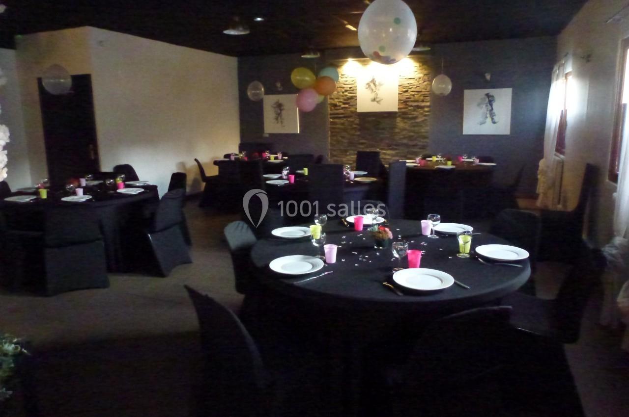 Salle décorée avec des tables rondes dressées, nappes noires, assiettes blanches et ballons suspendus au plafond.
