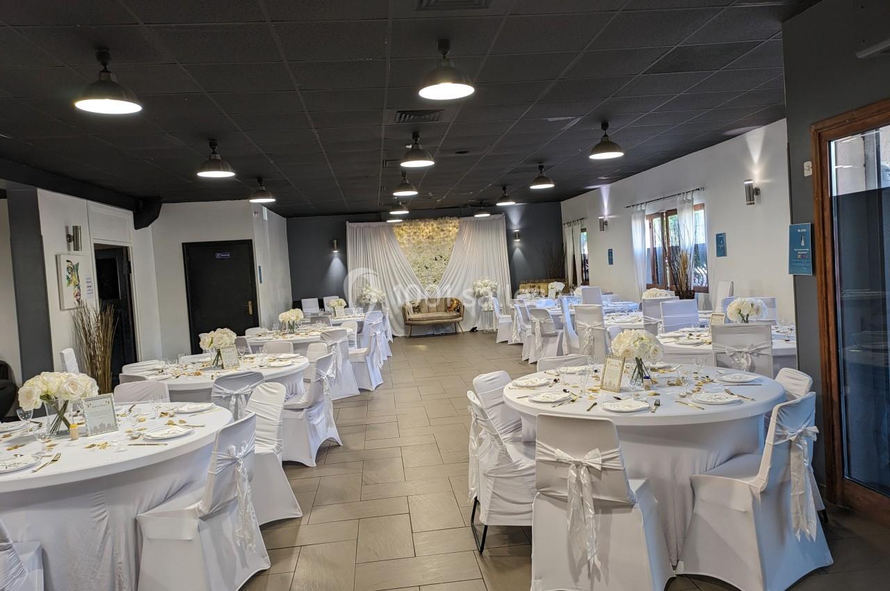 Salle de réception décorée avec des tables rondes couvertes de nappes blanches, chaises habillées et centre de table fleuris.