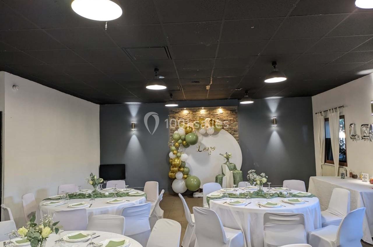 Salle décorée pour un événement avec tables rondes dressées, nappes blanches et arche de ballons blancs et dorés.