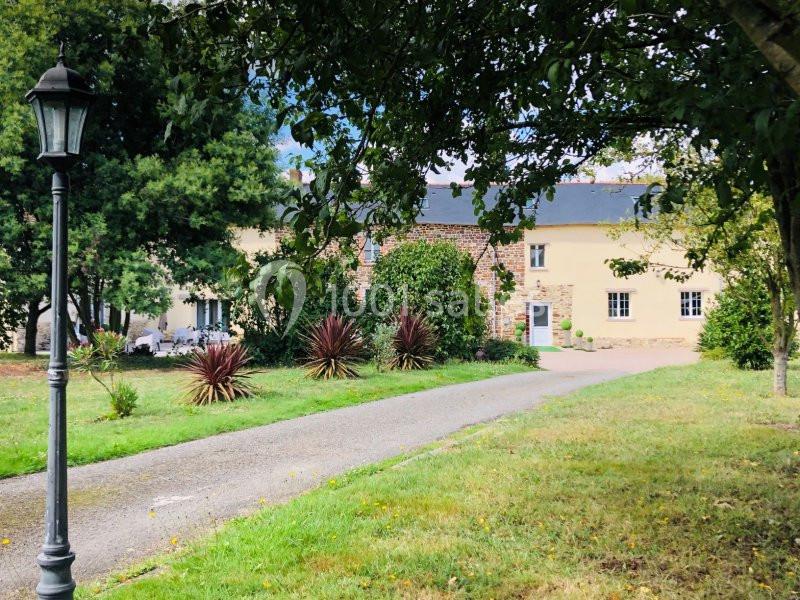 Allée bordée de pelouse menant à une maison en pierre et crépi jaune, entourée d'arbres et de plantes.