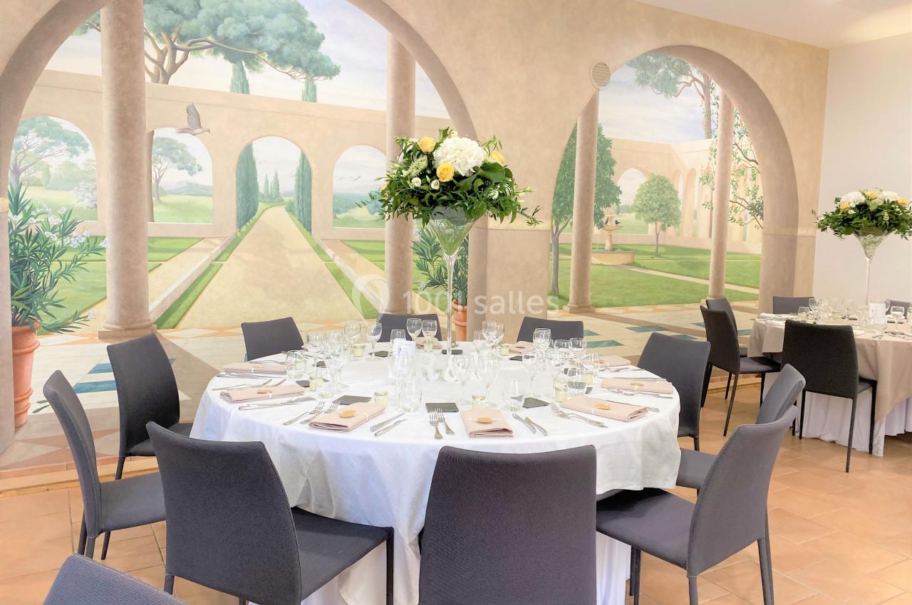 Salle de réception avec tables rondes dressées, chaises grises et fresque murale représentant un jardin méditerranéen.