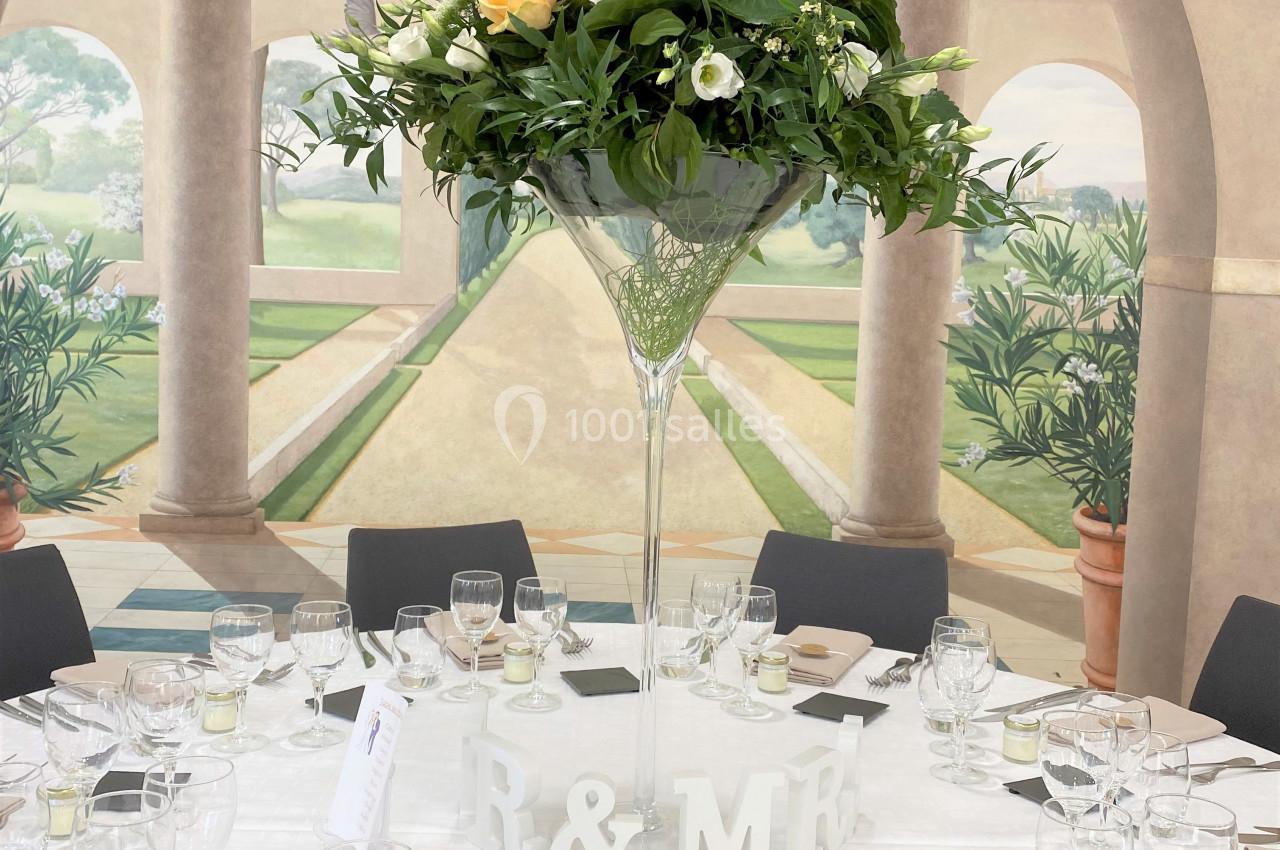 Centre de table floral avec roses jaunes et blanches dans un vase haut, entouré de vaisselle sur une table dressée.