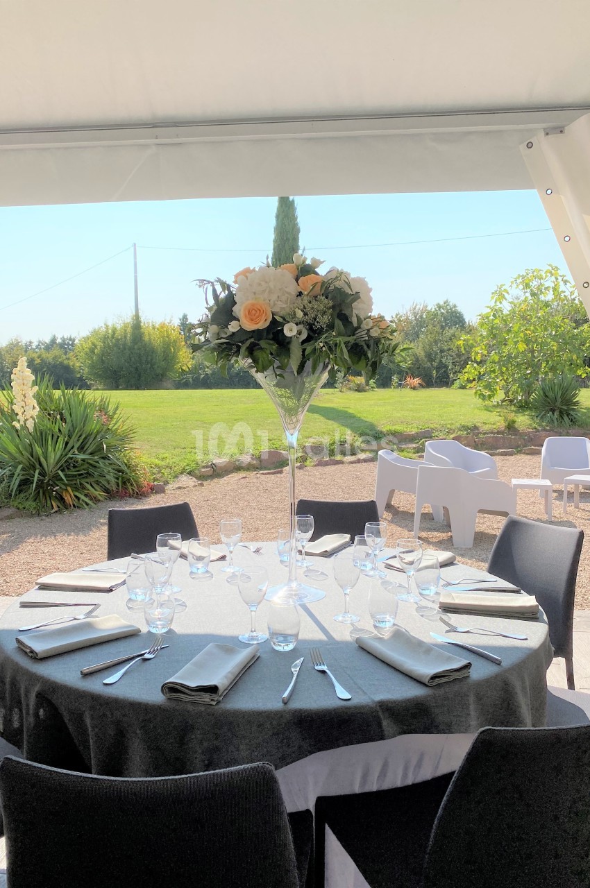 Table ronde dressée avec nappes grises, verres et couverts, décorée d'un centre floral, en extérieur sous une tente.
