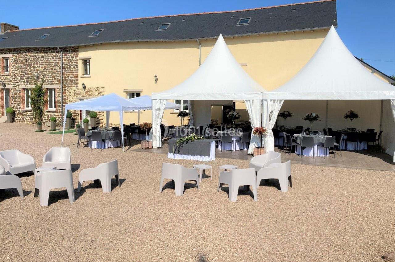 Cour extérieure avec barnums blancs, chaises et tables disposées pour un événement devant un bâtiment en pierre et crépi.