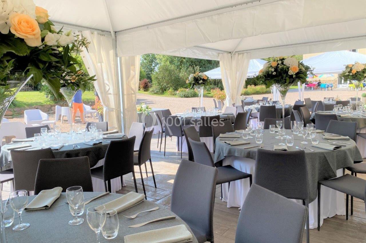 Salle de réception sous une tente blanche, avec tables dressées, nappes grises et décorations florales.