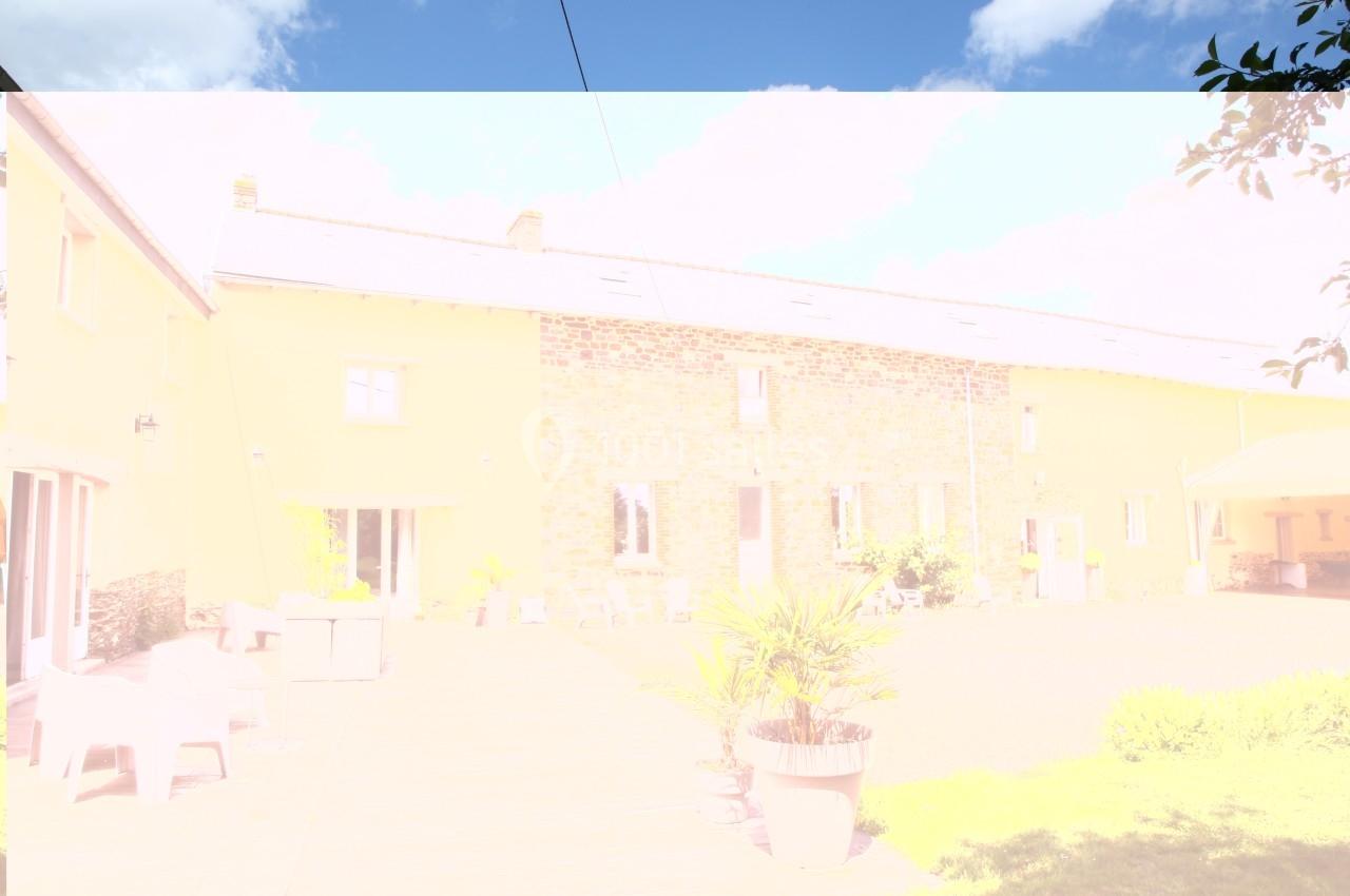 Façade d'une maison en pierre avec terrasse, mobilier de jardin et plantes, sous un ciel partiellement nuageux.