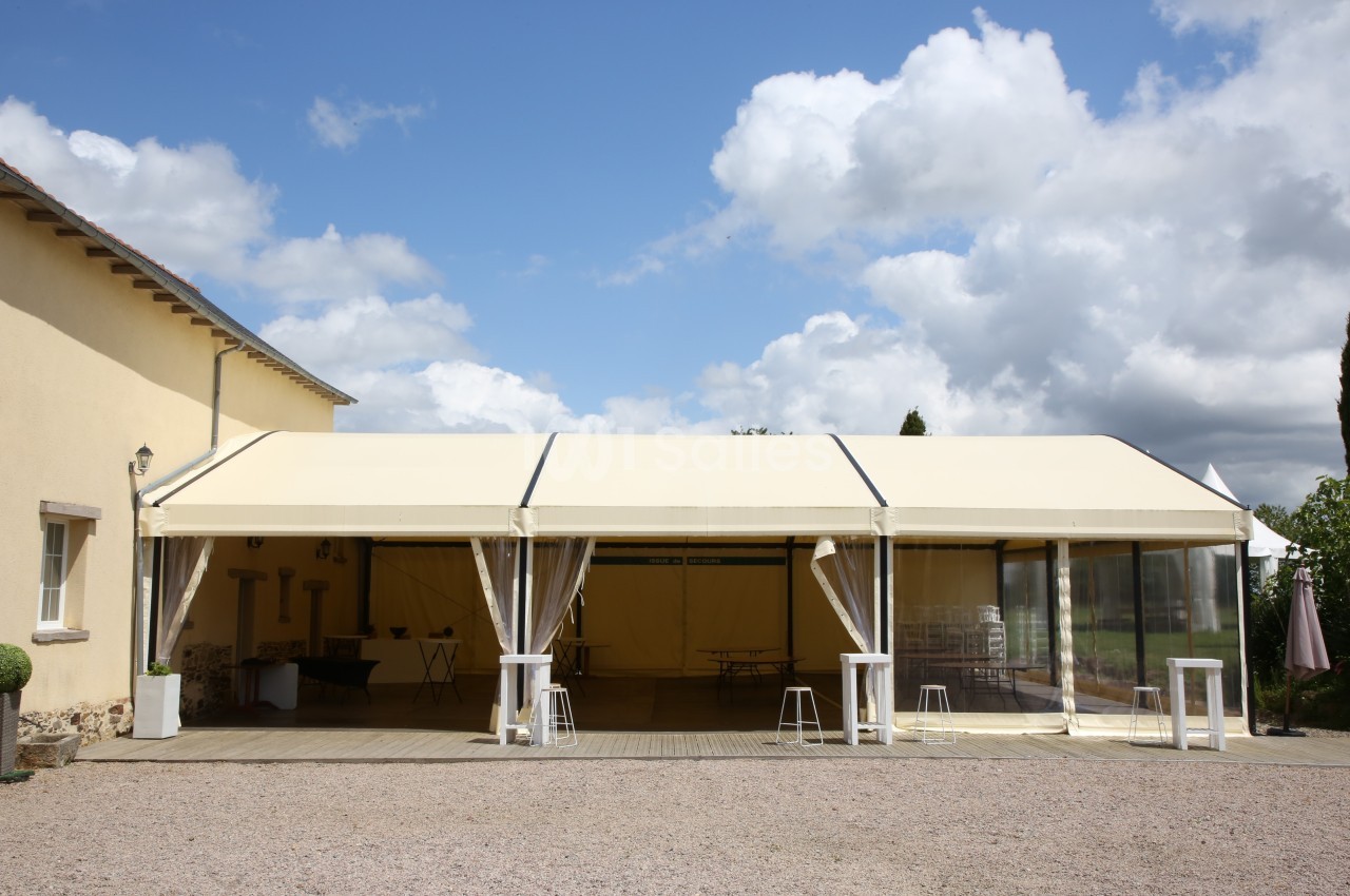 Tente beige installée sur une terrasse en bois, avec des tables et des chaises visibles à l'intérieur.