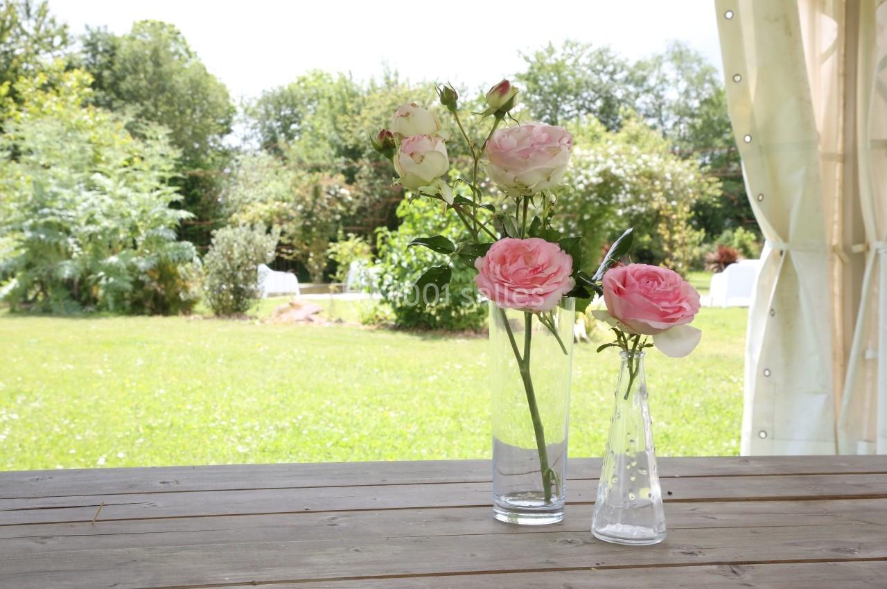 Deux vases en verre contenant des roses roses et blanches posés sur une table en bois, avec un jardin en arrière-plan.