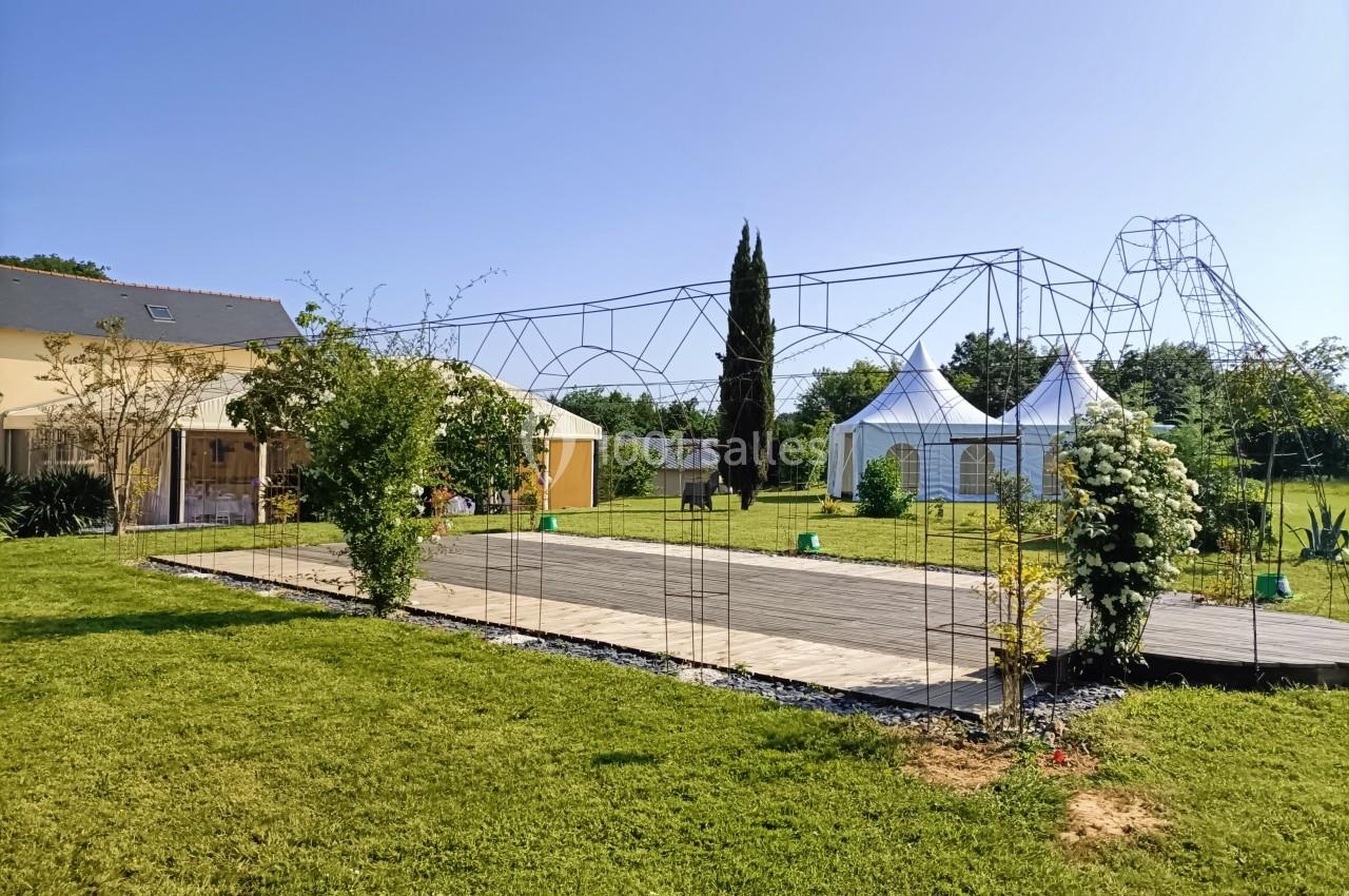 Jardin avec pelouse, pergola métallique, maison à gauche et tentes blanches en arrière-plan par temps ensoleillé.