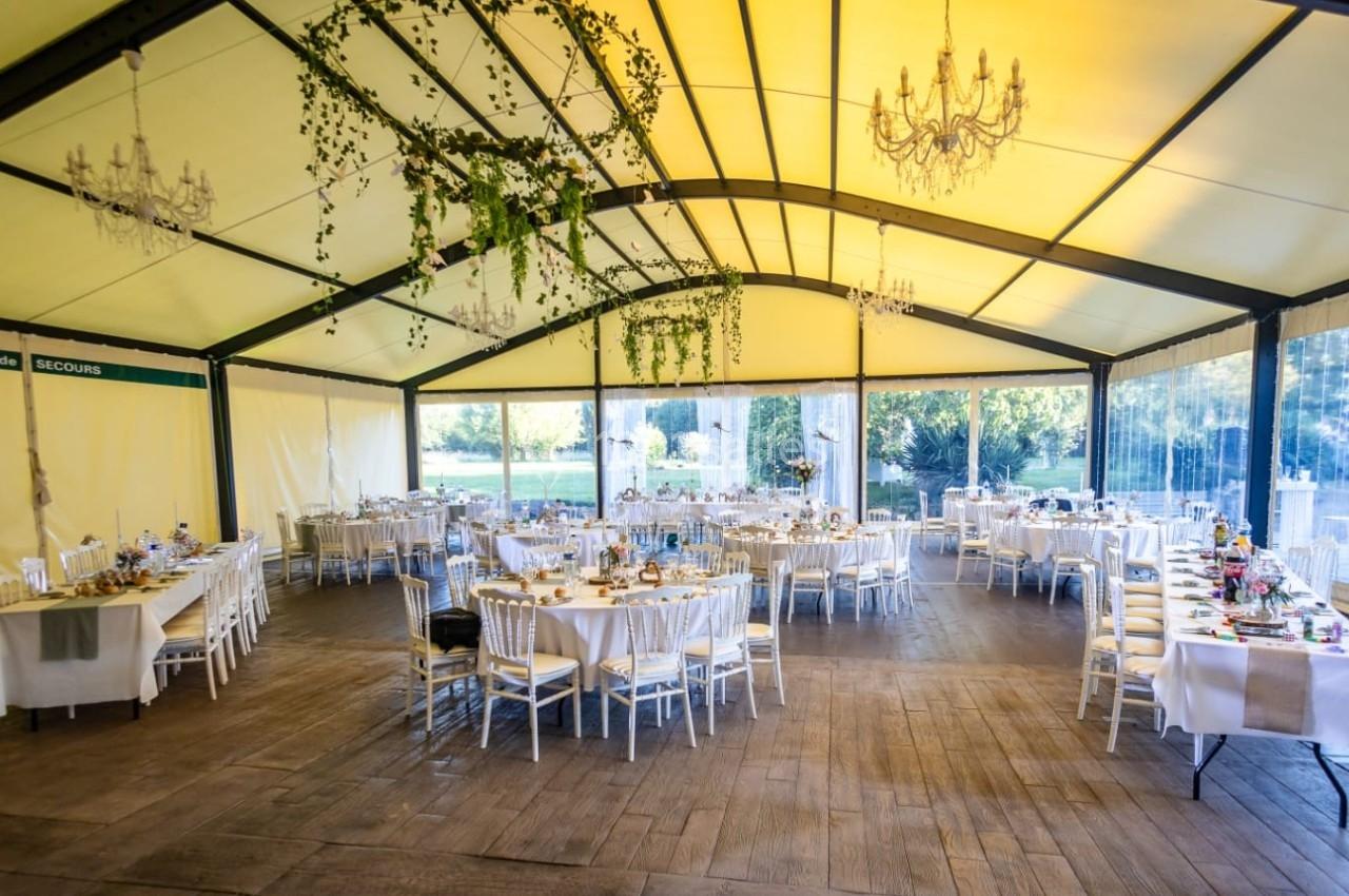 Salle de réception sous une tente avec tables décorées, chaises blanches et guirlandes végétales suspendues.