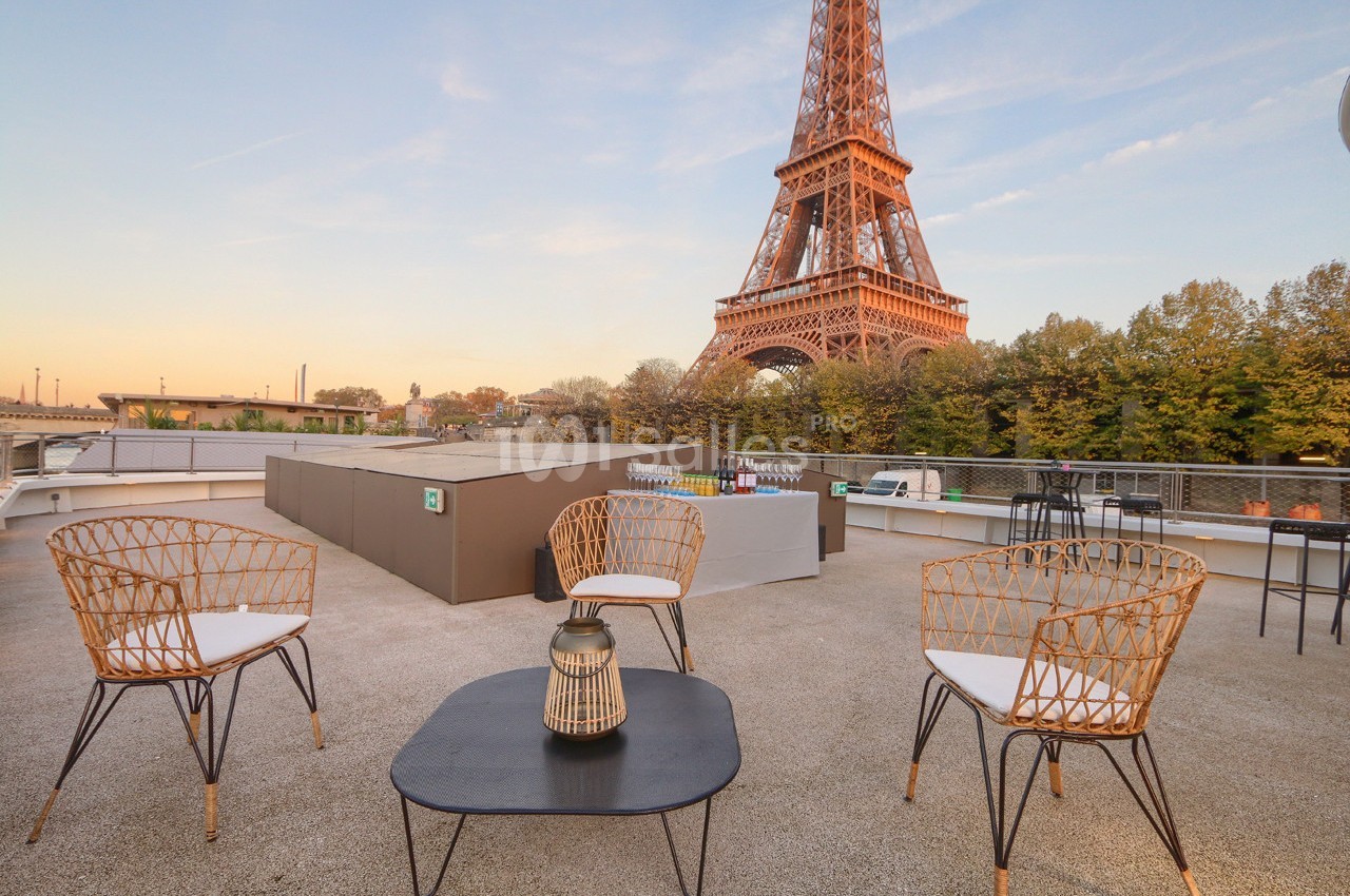 Terrasse avec mobilier en rotin, vue sur la tour Eiffel au coucher du soleil et arbres en arrière-plan.