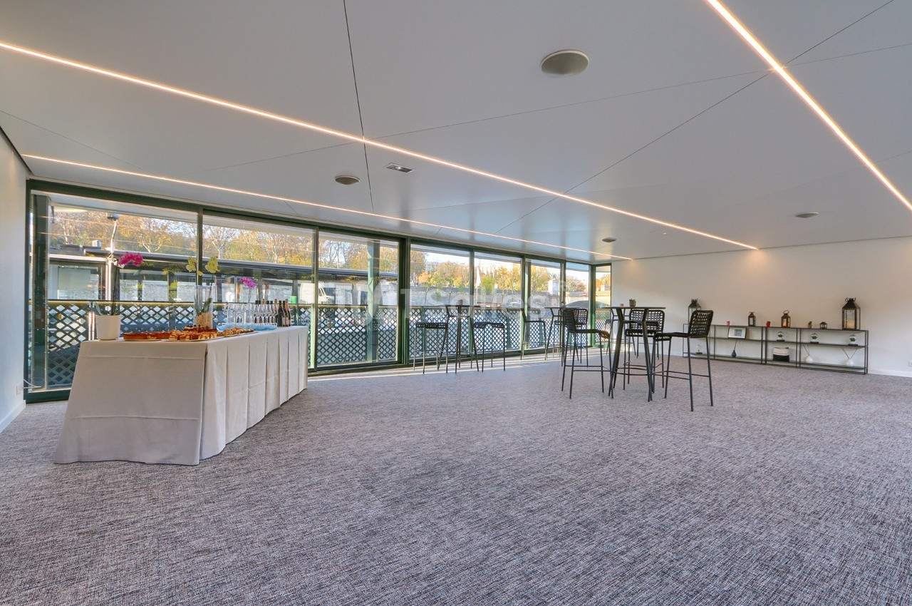 Salle lumineuse avec moquette grise, grandes baies vitrées, table de buffet et chaises hautes.