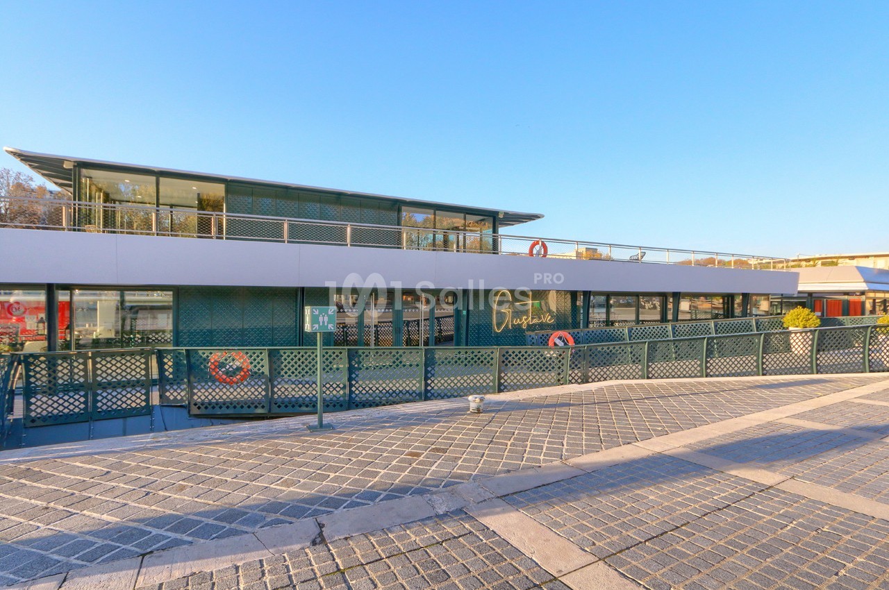 Bâtiment moderne flottant avec façade vitrée, entouré d'une passerelle et situé sur un quai pavé.