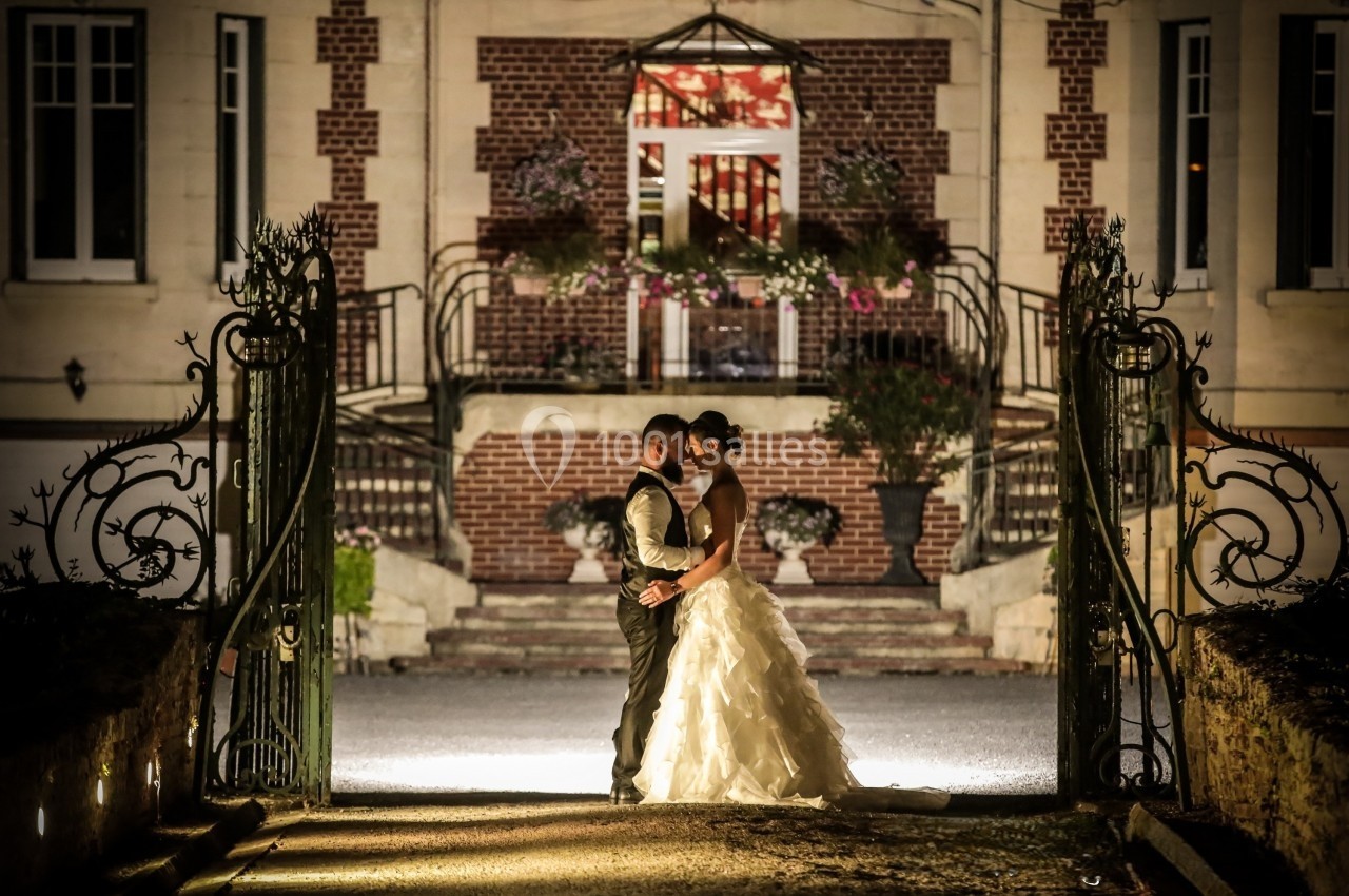 Un couple en tenue de mariage pose devant une entrée illuminée d'un bâtiment en briques et pierre.