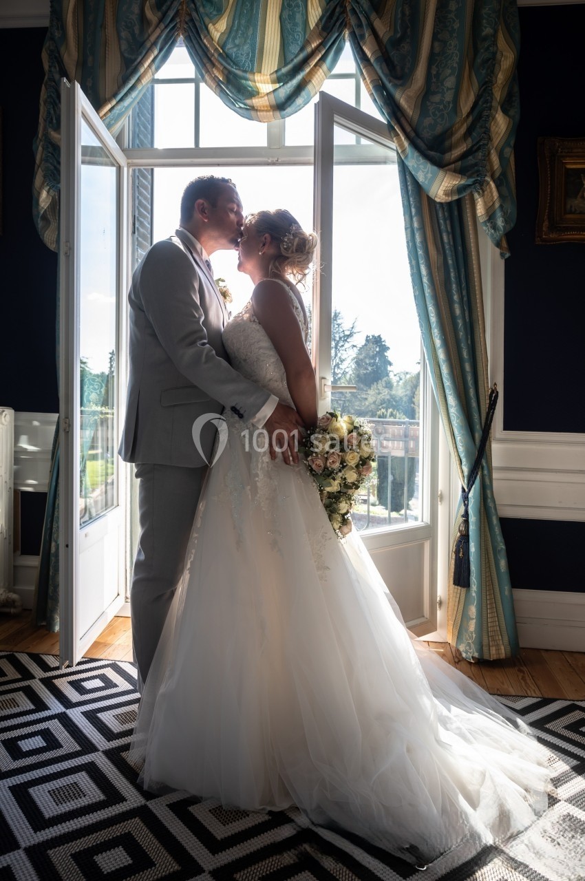 Un couple de mariés s'embrasse devant une porte-fenêtre laissant entrer la lumière, dans une pièce élégante.