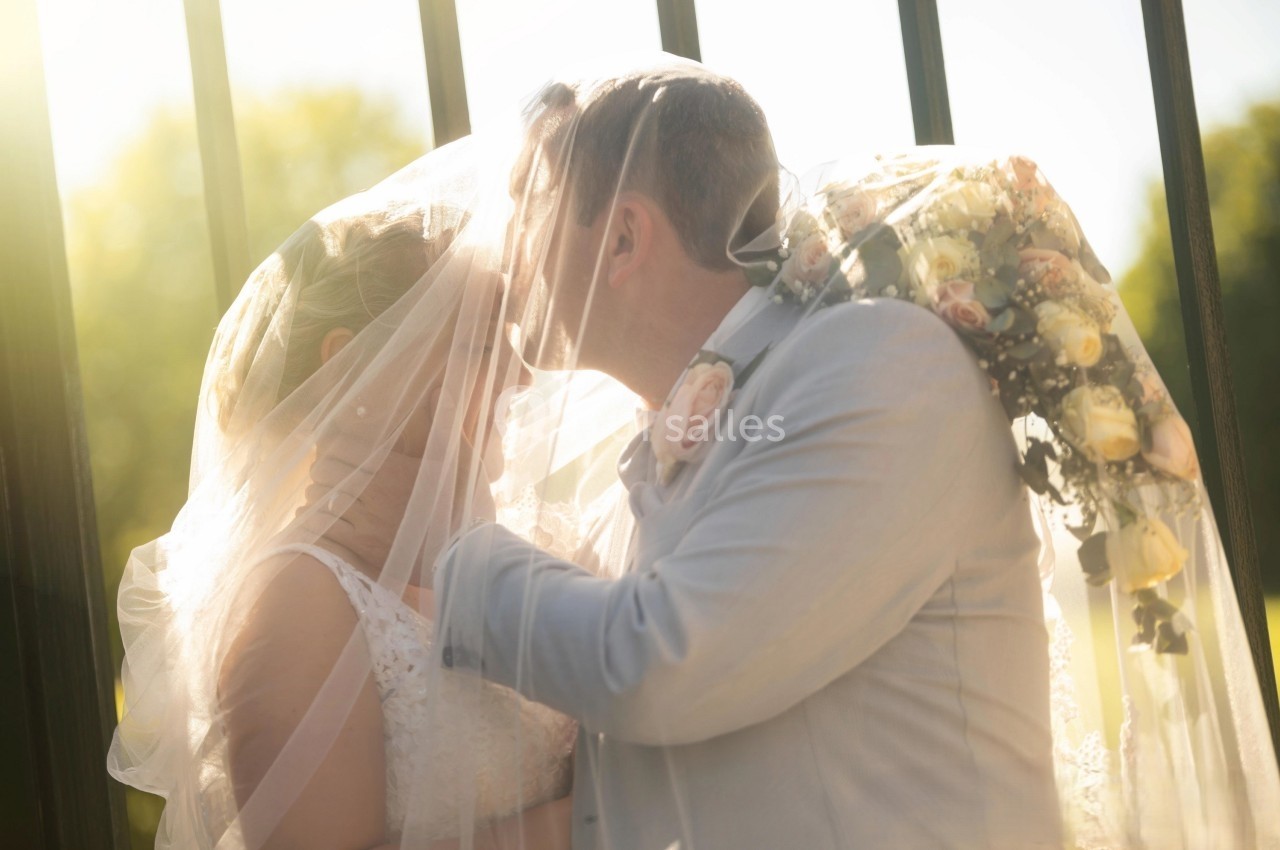 Un couple de mariés s'embrasse tendrement sous un voile, avec un bouquet de fleurs pastel en arrière-plan.