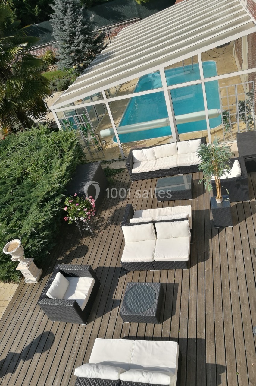 Terrasse en bois avec salon de jardin, plantes en pot et vue sur une véranda abritant une piscine.