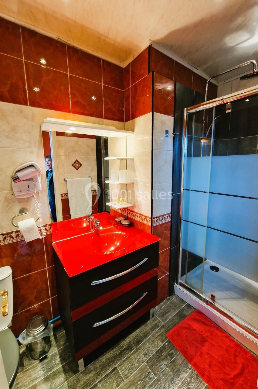 Salle de bain avec lavabo rouge, miroir éclairé, douche vitrée, carrelage beige et rouge, et accessoires muraux.