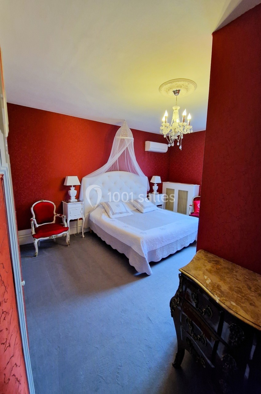 Chambre élégante avec murs rouges, lit double à baldaquin blanc, mobilier classique et lustre au plafond.