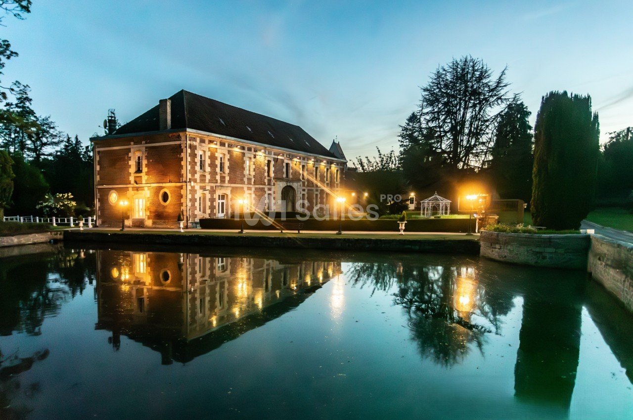 Bâtiment historique en pierre éclairé, reflété dans un plan d'eau au crépuscule, entouré de verdure.