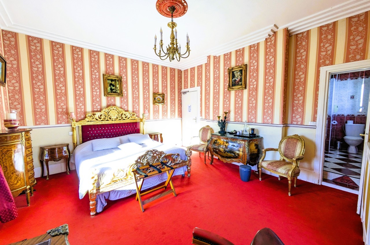 Chambre avec lit double, mobilier ancien, tapis rouge, murs rayés et vue sur une salle de bain avec carrelage noir et blanc.