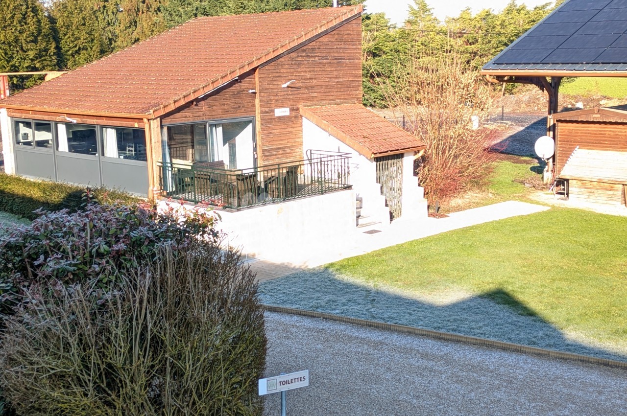 Bâtiment en bois avec terrasse, entouré d'un jardin, chemin gravillonné et panneau indiquant des toilettes au premier plan.
