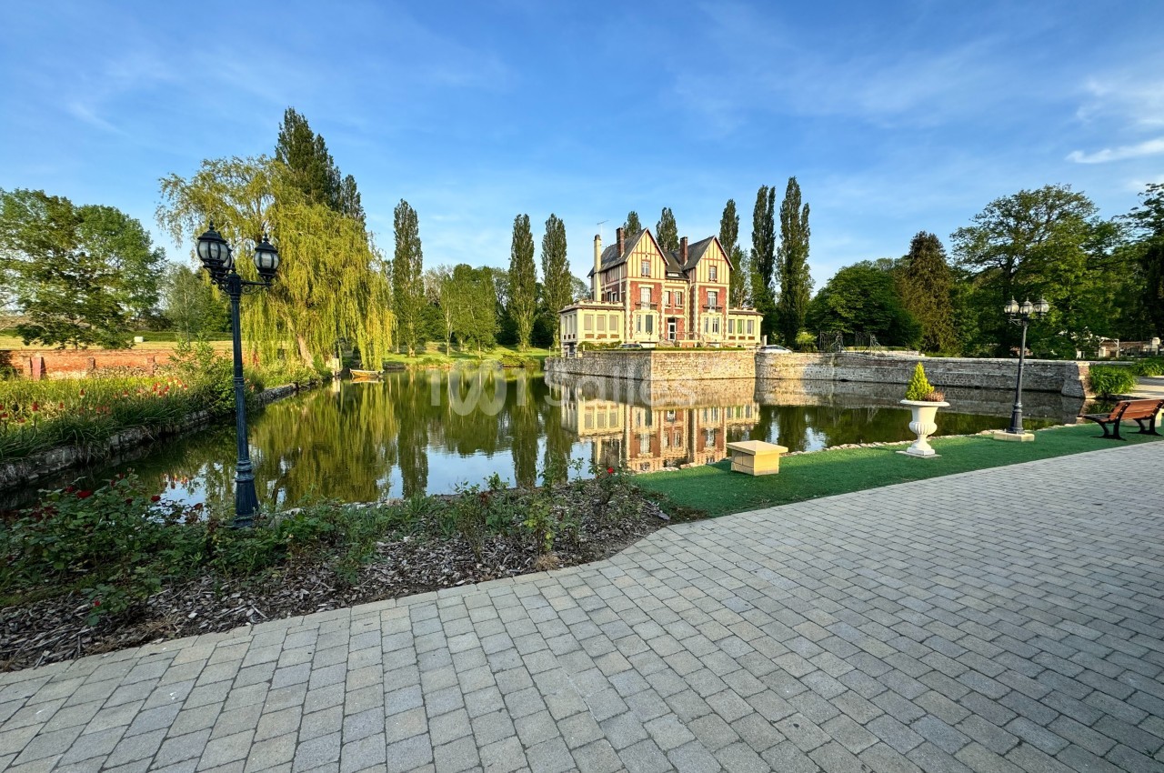 Manoir entouré d'arbres, reflet dans un étang, bordé d'une allée pavée et d'un jardin paysager.