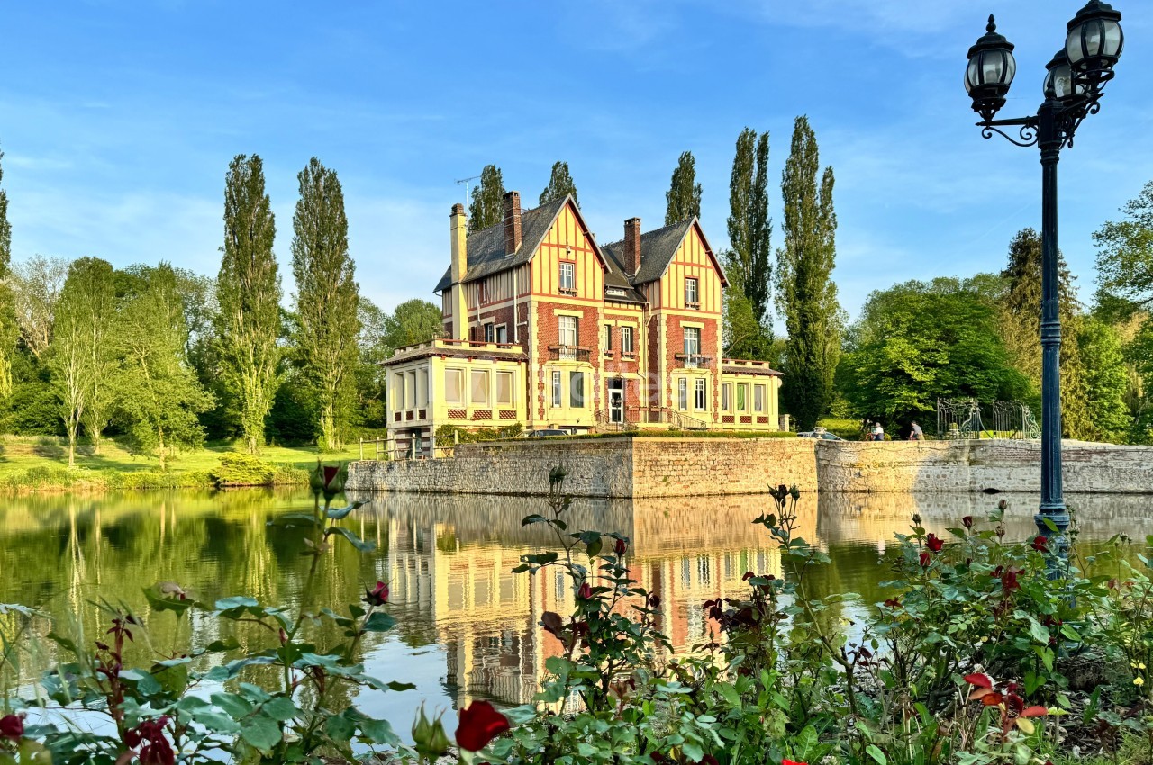 Manoir en briques rouges entouré d'arbres, reflété dans un étang avec des rosiers au premier plan.