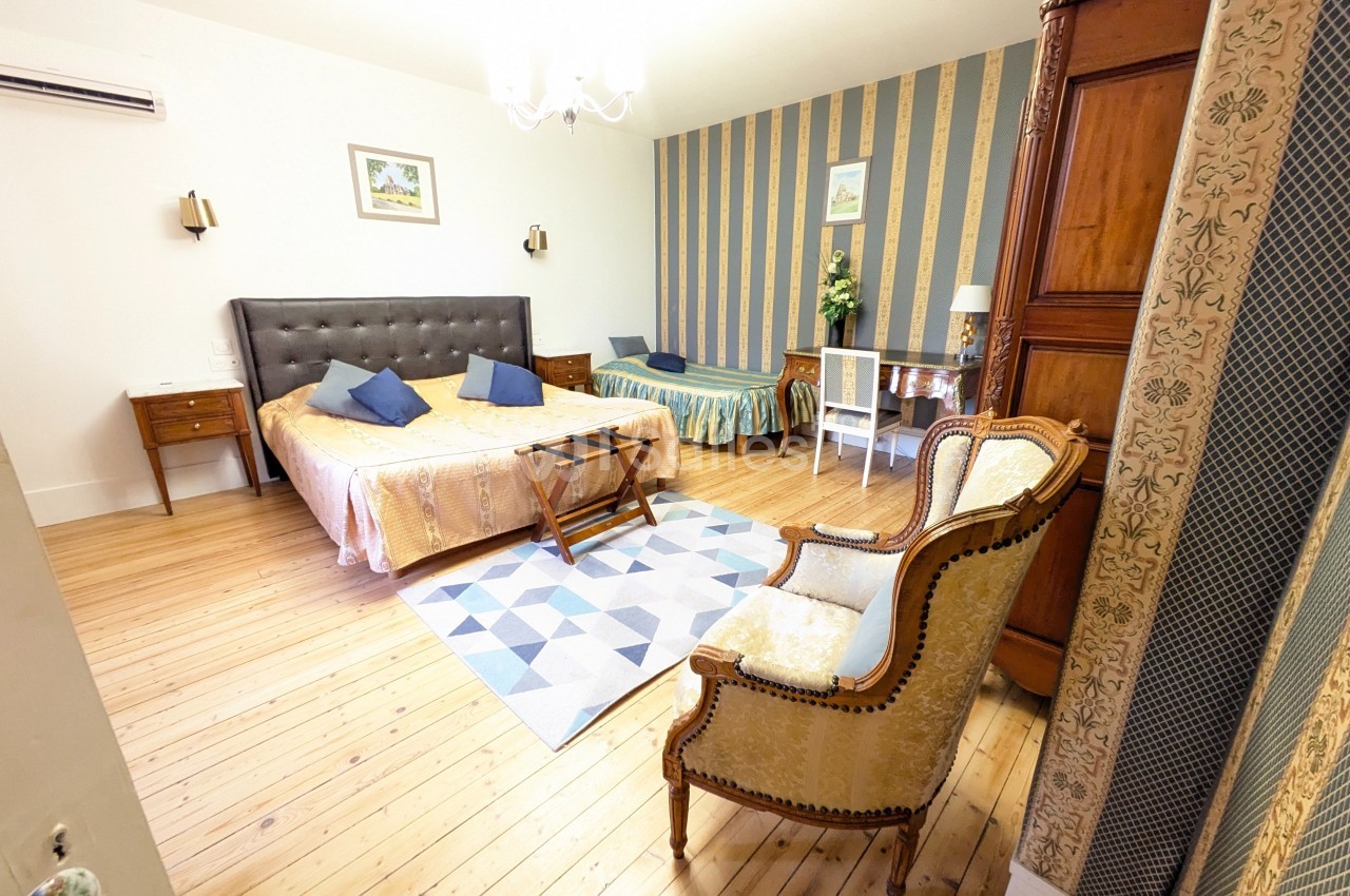 Chambre spacieuse avec lit double, lit simple, fauteuil, armoire en bois, et décoration classique sur parquet clair.