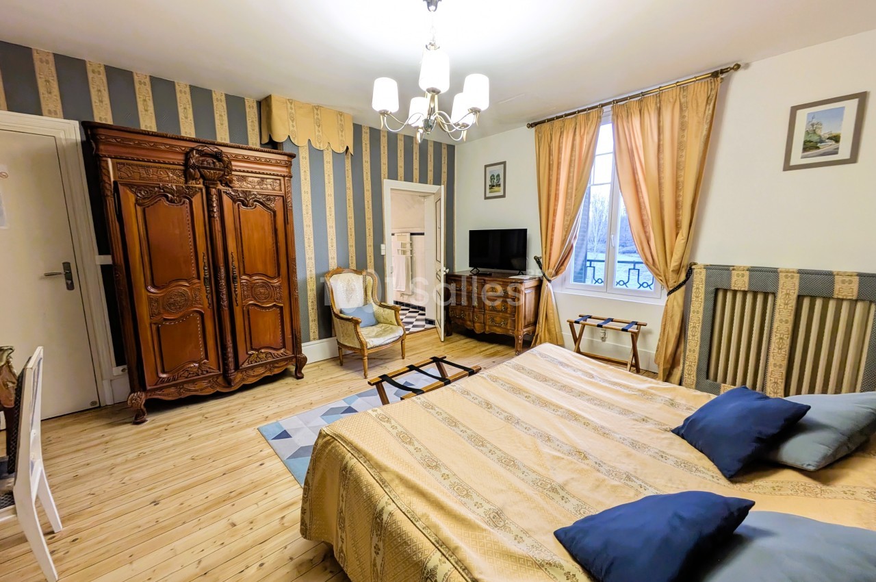 Chambre lumineuse avec lit double, armoire en bois sculpté, fauteuil, télévision et rideaux dorés.