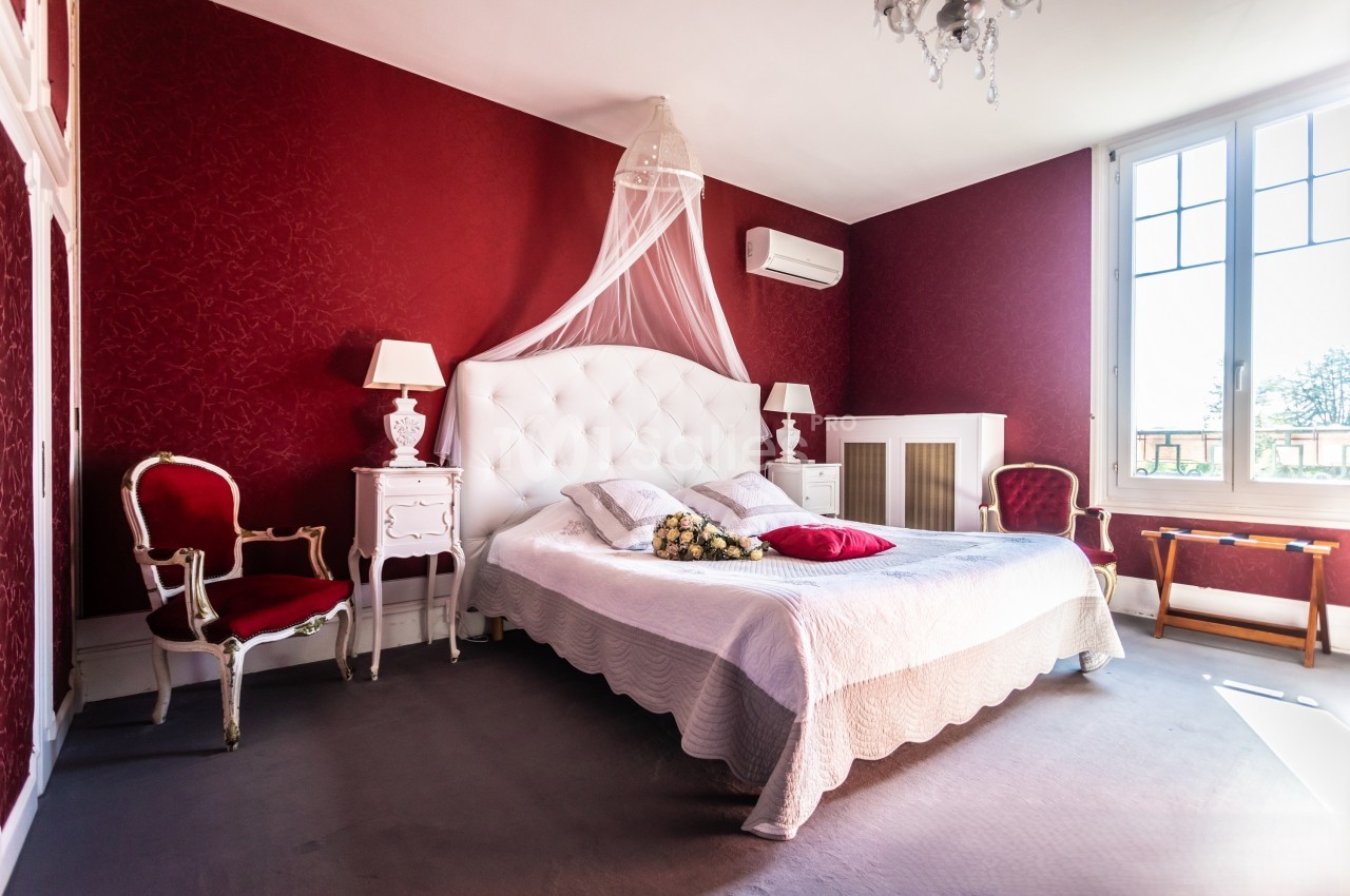 Chambre élégante avec murs rouges, lit double à tête capitonnée blanche, mobilier classique et lumière naturelle.
