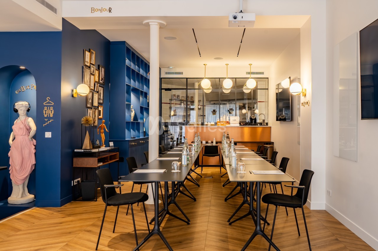 Salle de réunion moderne avec tables alignées, chaises noires, éclairage suspendu et décoration artistique.