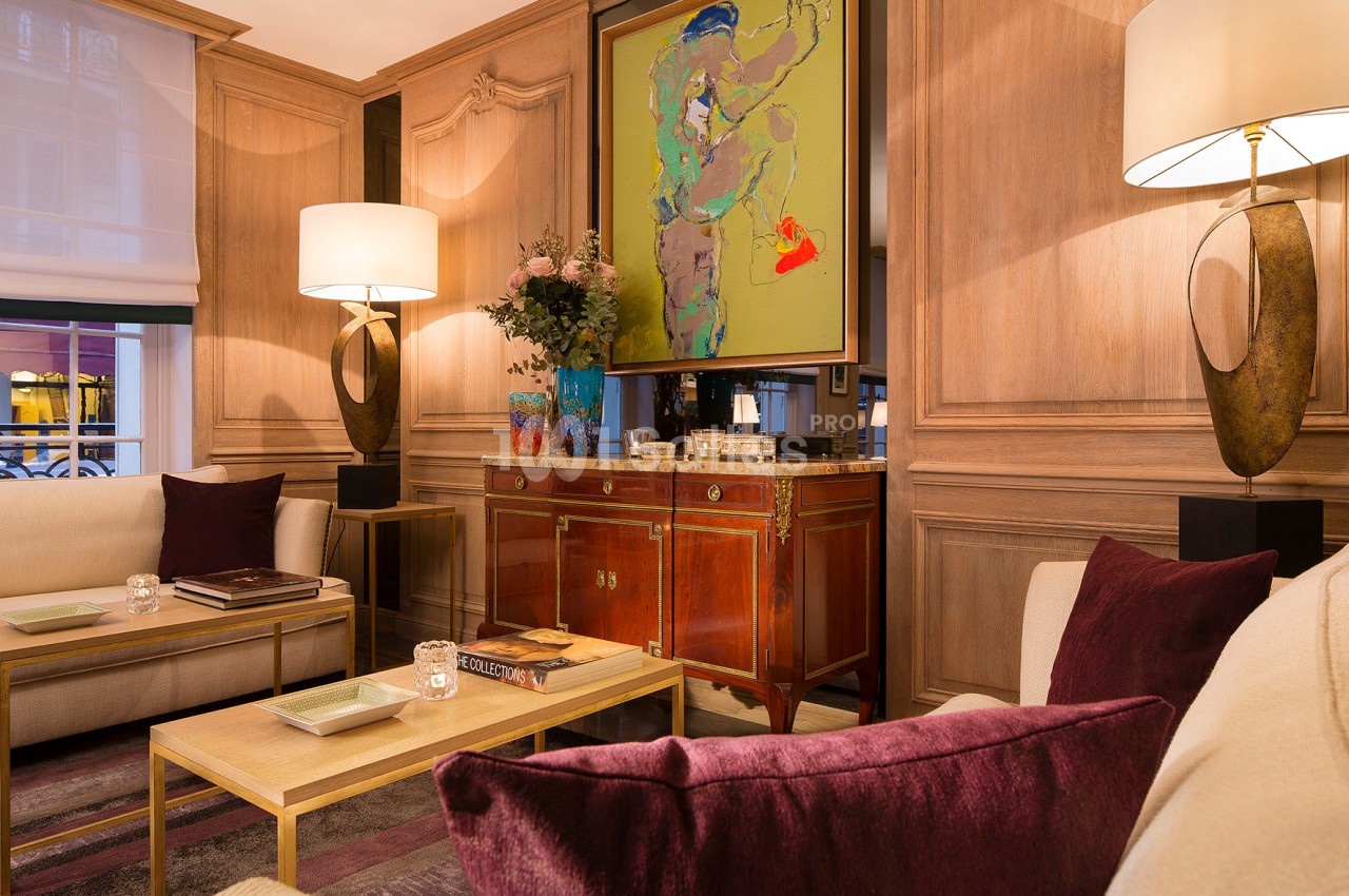Salon élégant avec boiseries, meubles classiques, tableau coloré, lampes modernes et coussins violets sur les canapés.