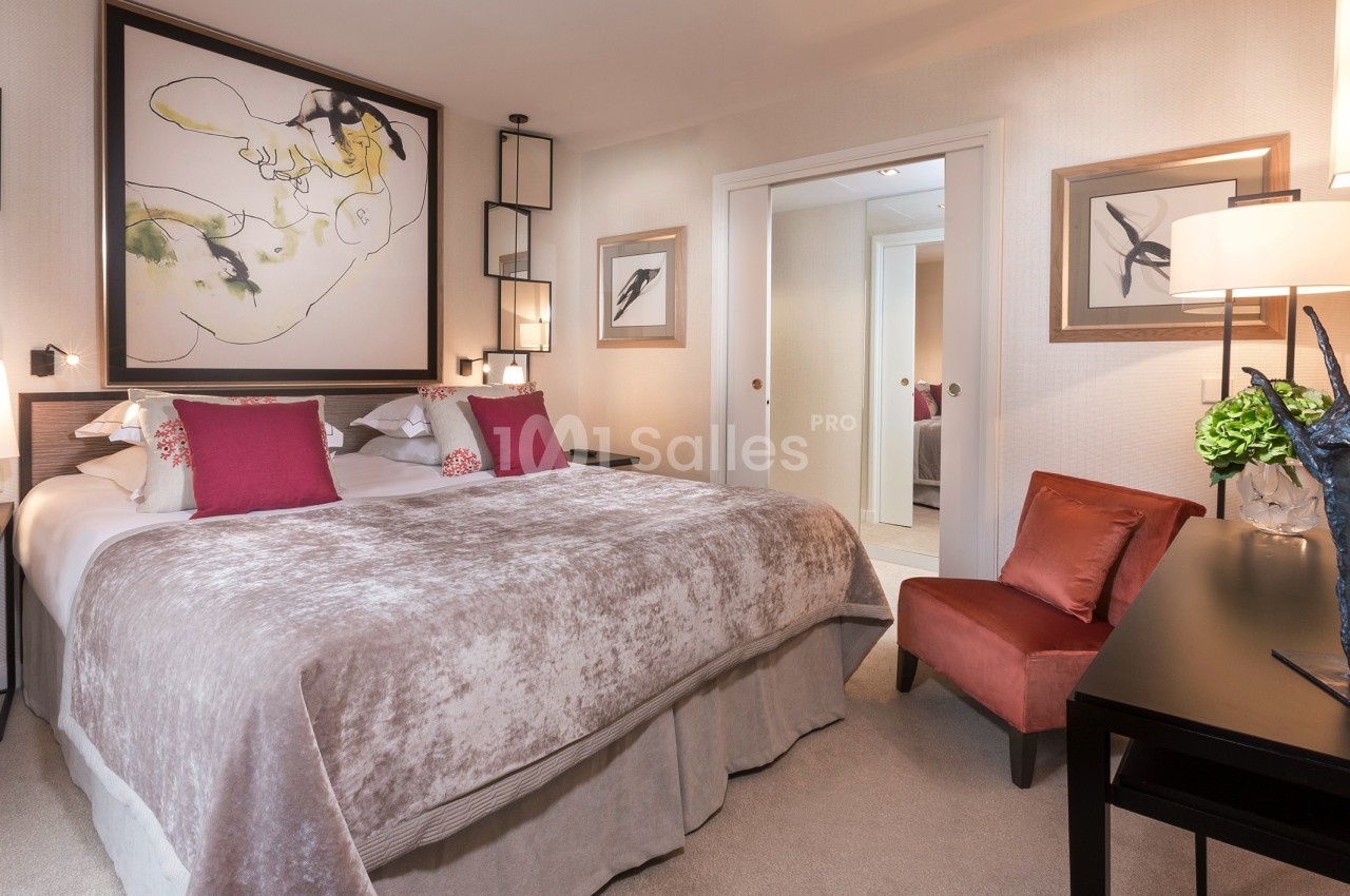Chambre élégante avec un grand lit, des coussins colorés, des œuvres d'art murales et un fauteuil près d'une table noire.