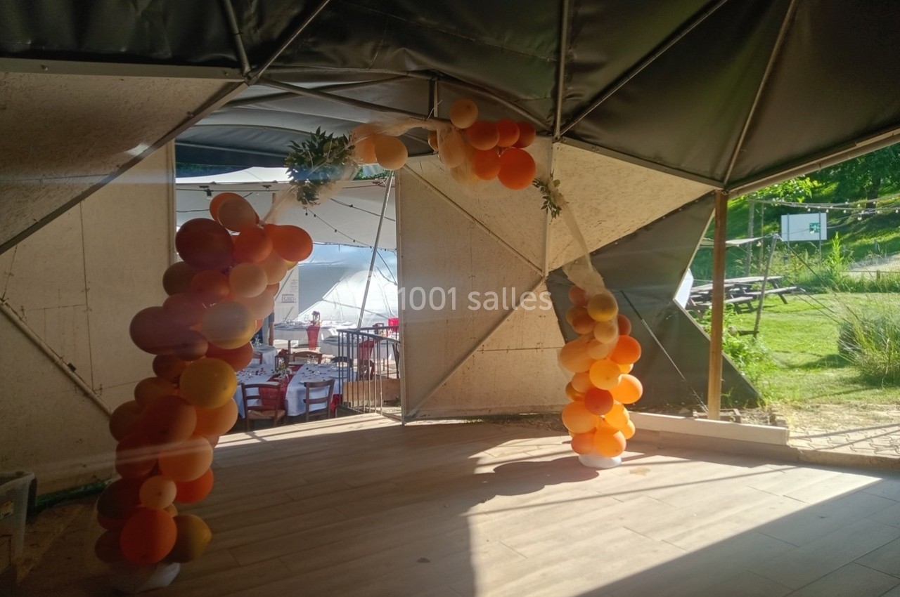 Arche décorative en ballons orange et jaunes dans un espace extérieur ensoleillé avec tables et verdure en arrière-plan.