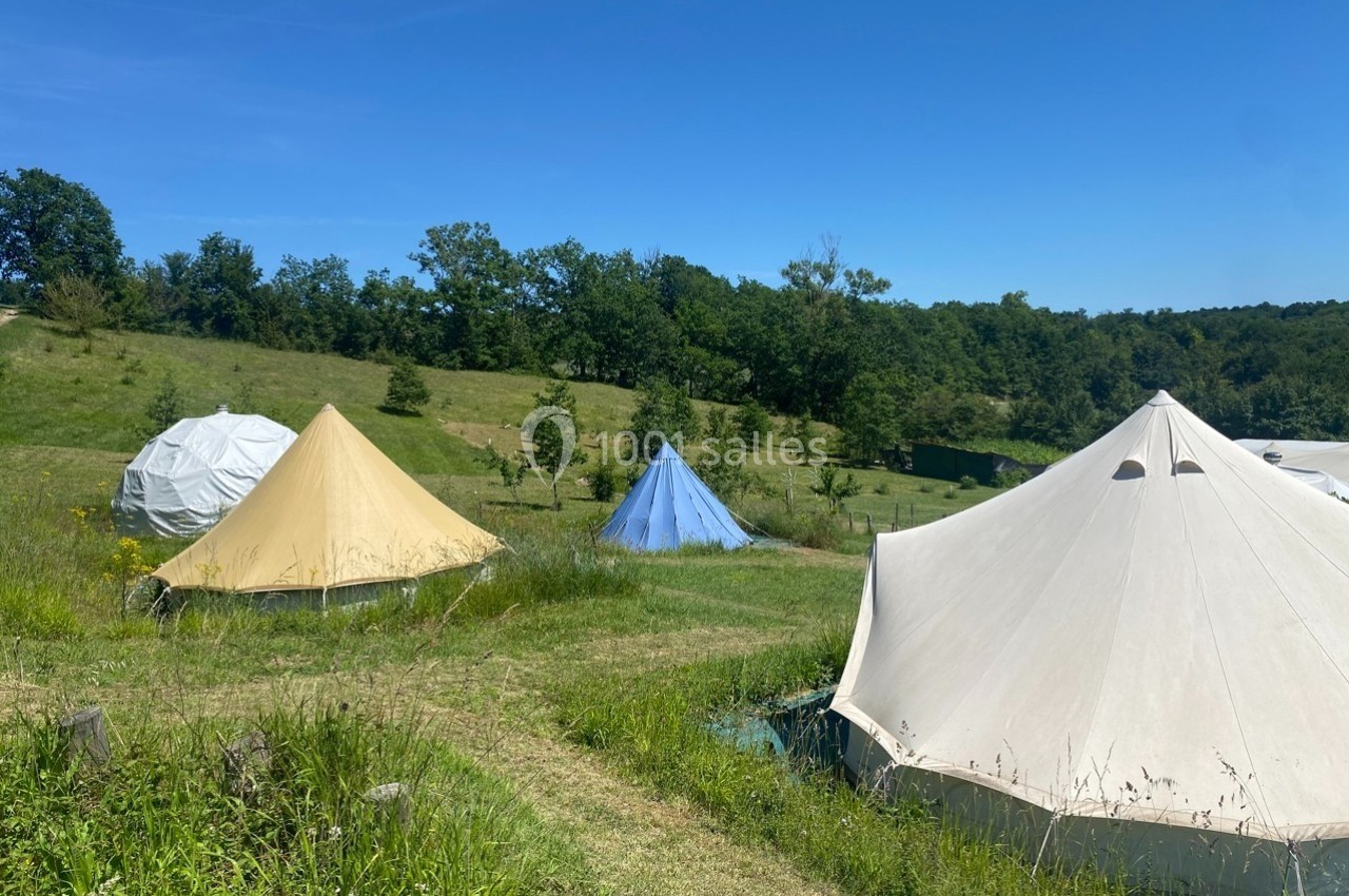 Tentes de camping installées sur une prairie verdoyante sous un ciel bleu, entourées d'arbres et de collines.