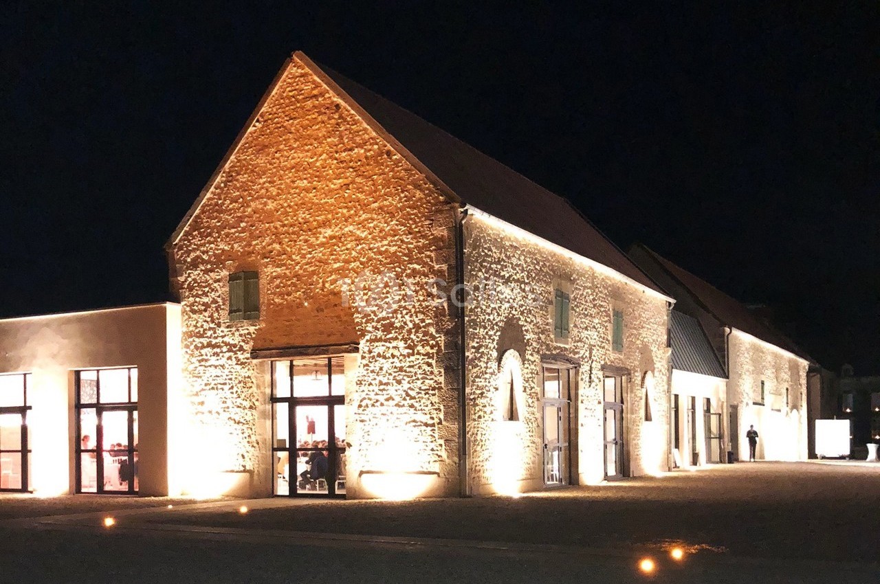 Bâtiment en pierre éclairé la nuit, avec grandes fenêtres illuminées et une cour devant.