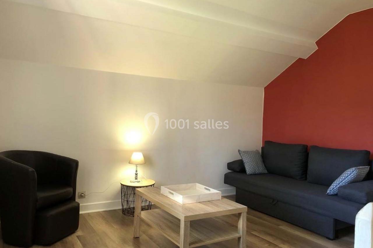 Salon avec un canapé gris, un fauteuil noir, une table basse en bois clair et un mur rouge en arrière-plan.