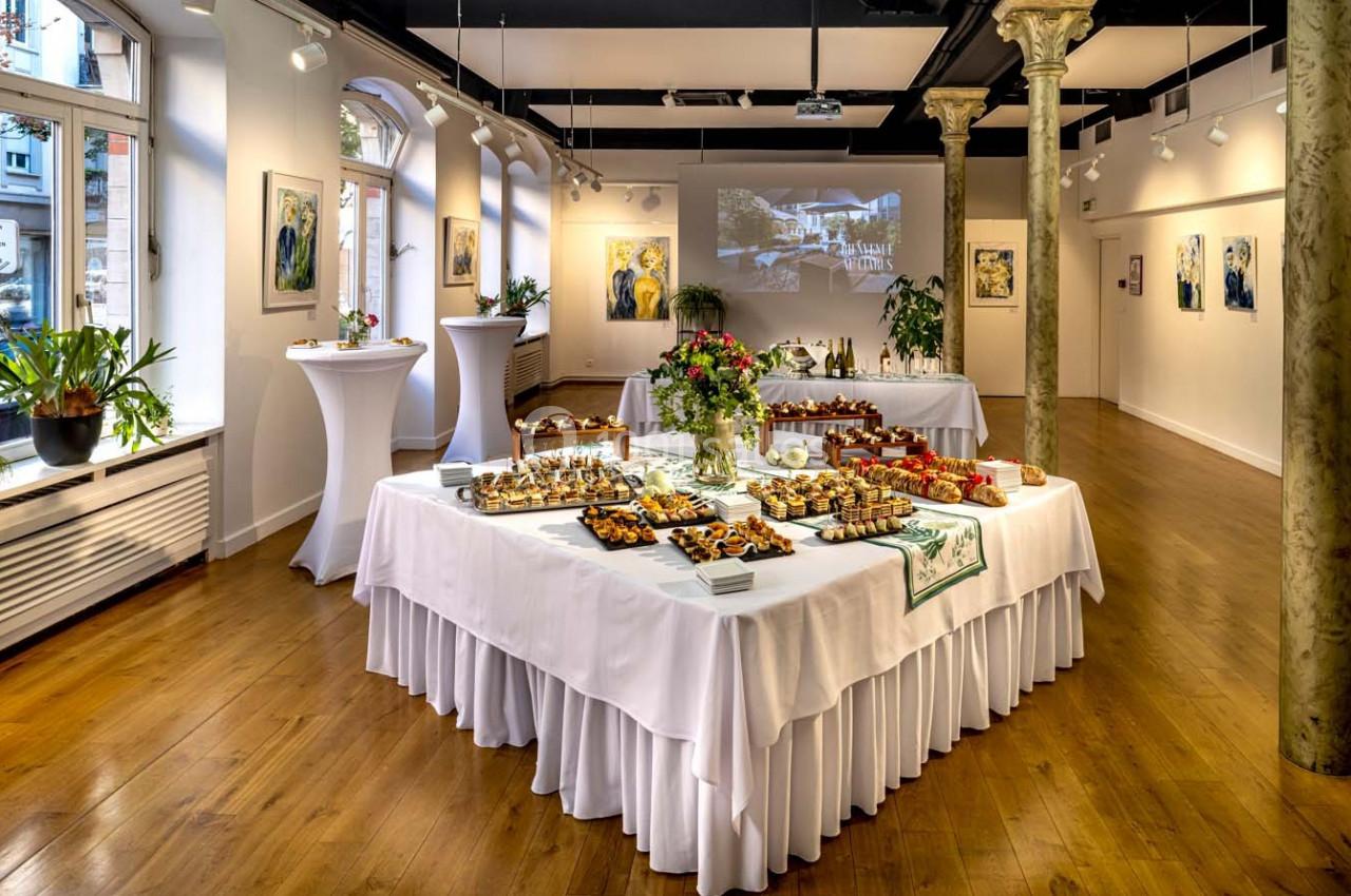 Salle lumineuse avec parquet, tables dressées pour un buffet, décorée de plantes et de tableaux accrochés aux murs.
