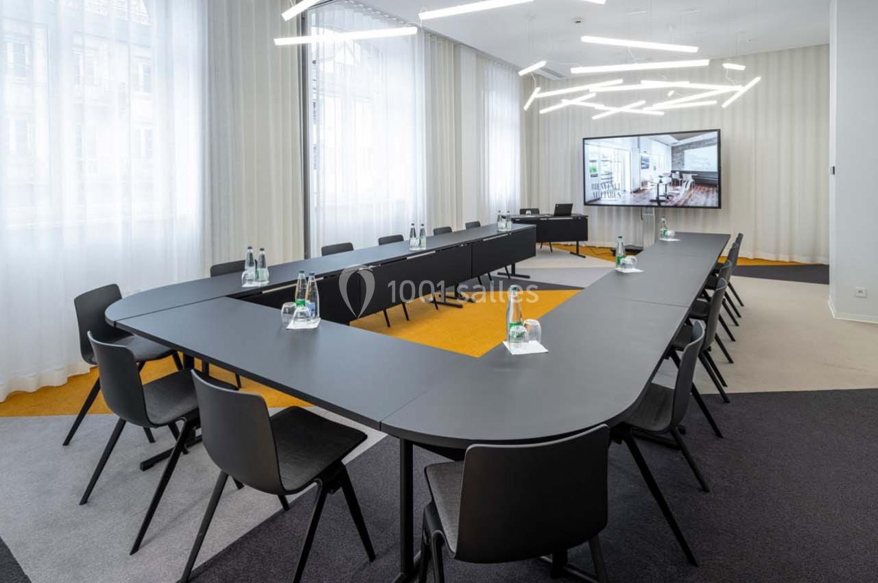Salle de réunion moderne avec tables en U, chaises noires, bouteilles d'eau, écran et éclairage suspendu.