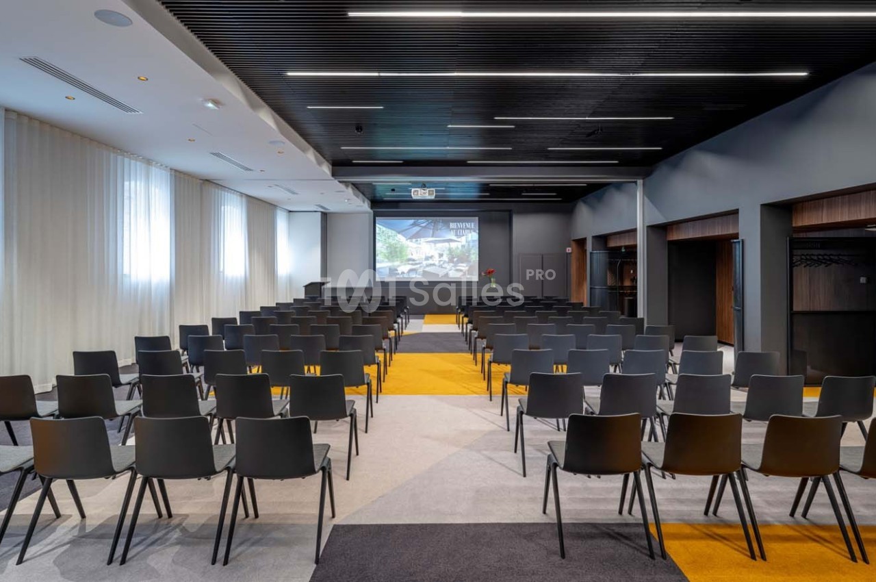 Salle de conférence moderne avec rangées de chaises noires, écran de projection allumé et éclairage naturel.