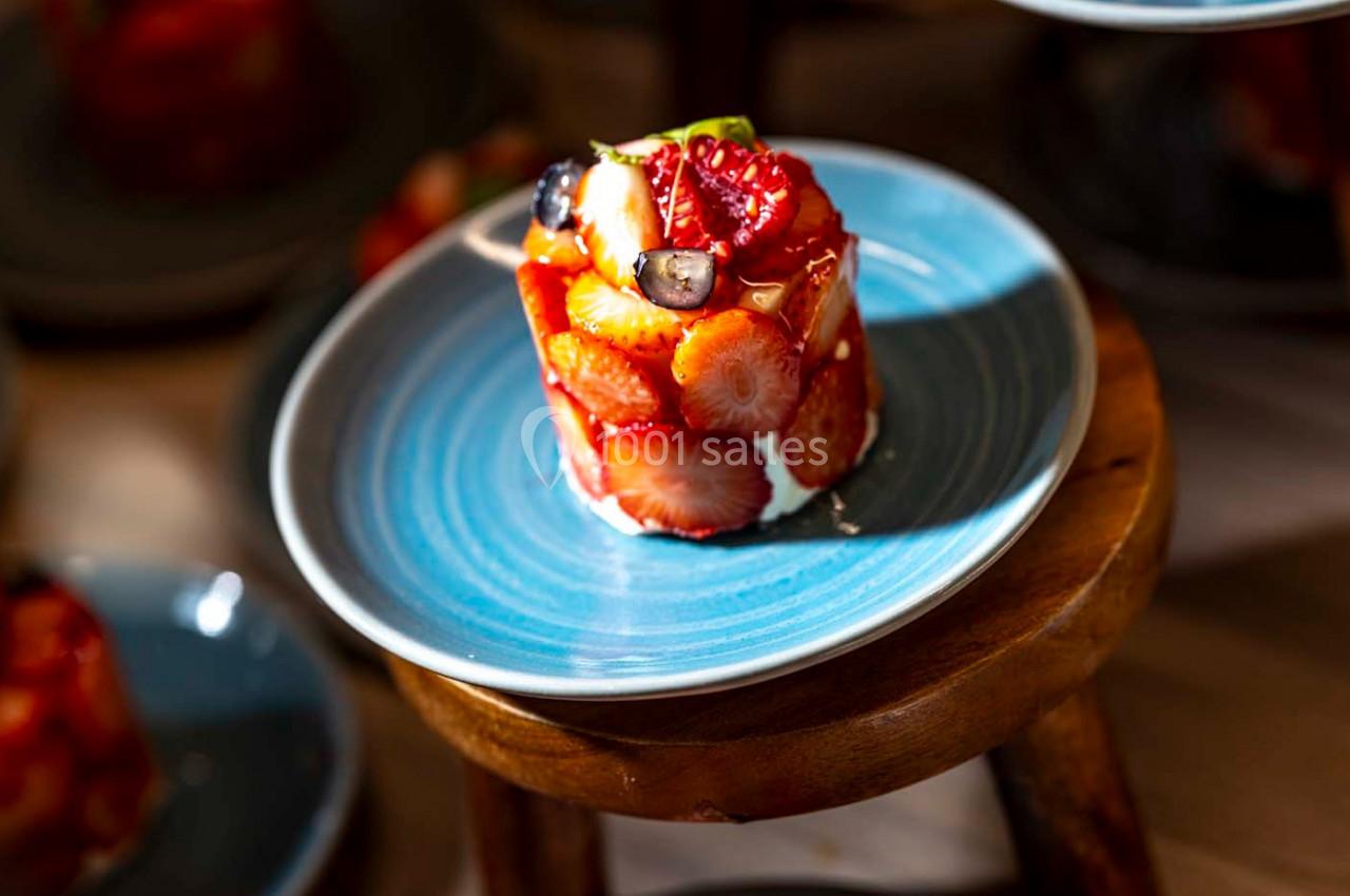 Dessert composé de fraises, fruits rouges et crème, présenté sur une assiette bleue posée sur un tabouret en bois.
