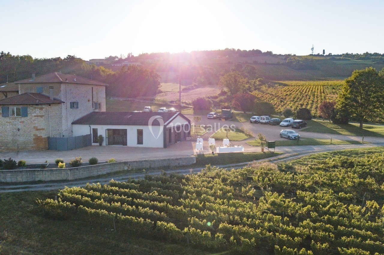 Vue aérienne d'un domaine viticole avec bâtiments, vignobles et voitures stationnées sous un soleil couchant.