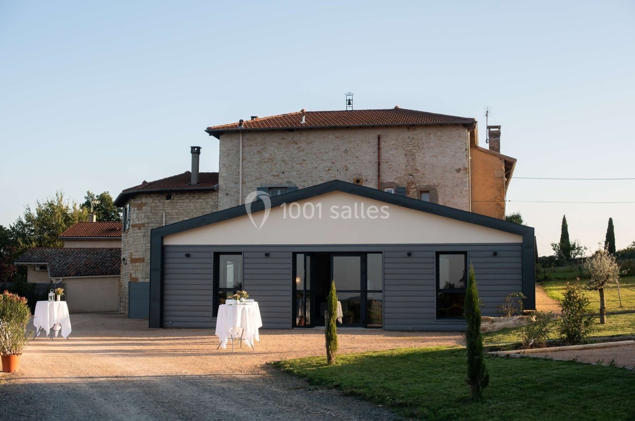 Bâtiment en pierre avec une extension moderne, tables dressées à l'extérieur sur une allée gravillonnée.