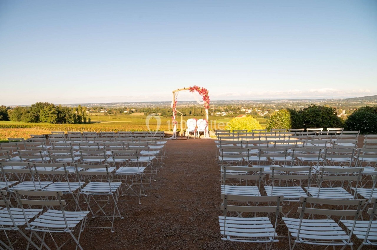 Chaises blanches alignées en extérieur face à une arche décorée de fleurs, avec vue sur un paysage de campagne.