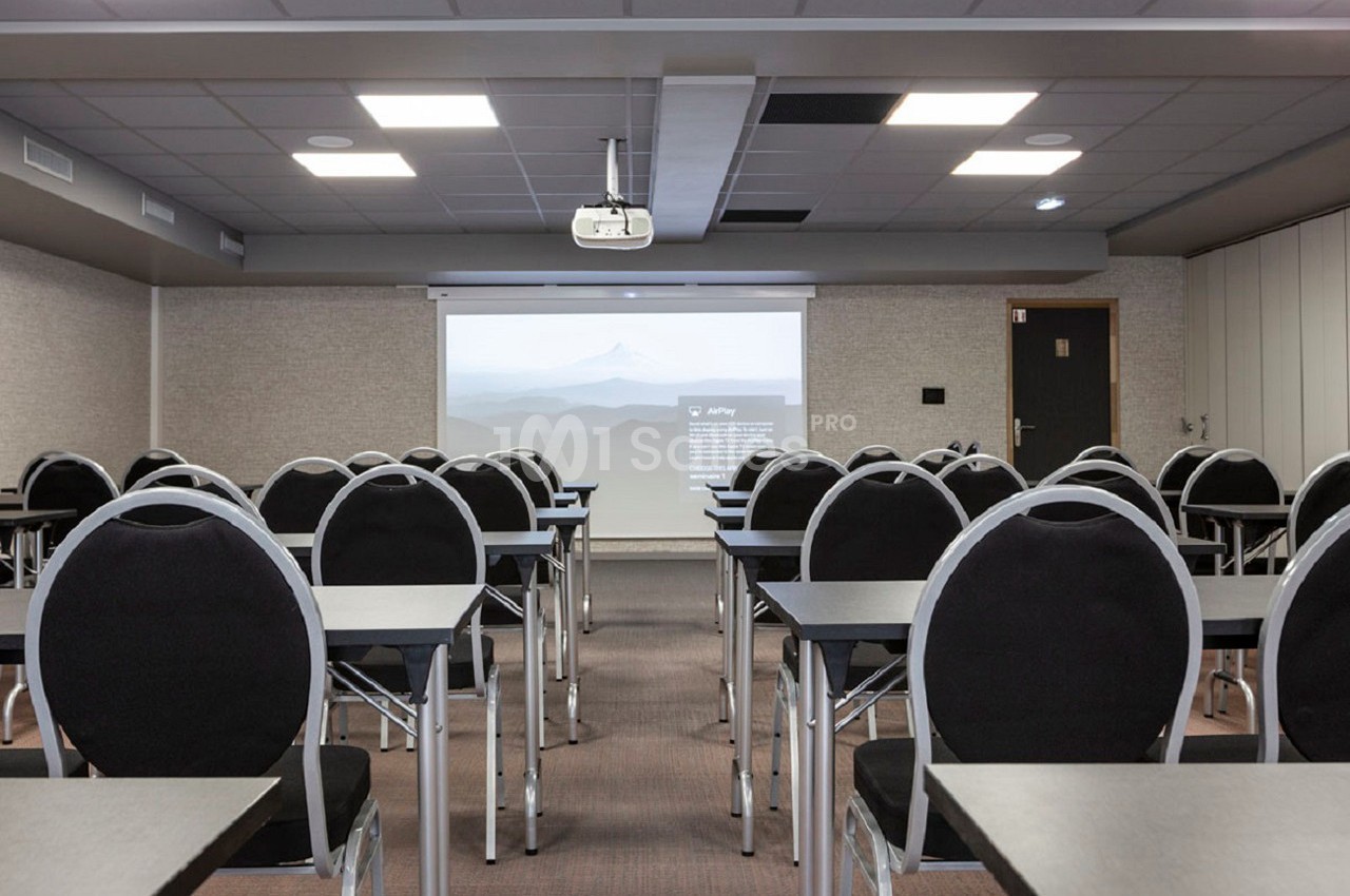 Salle de conférence vide avec des rangées de chaises noires et des tables face à un écran de projection allumé.
