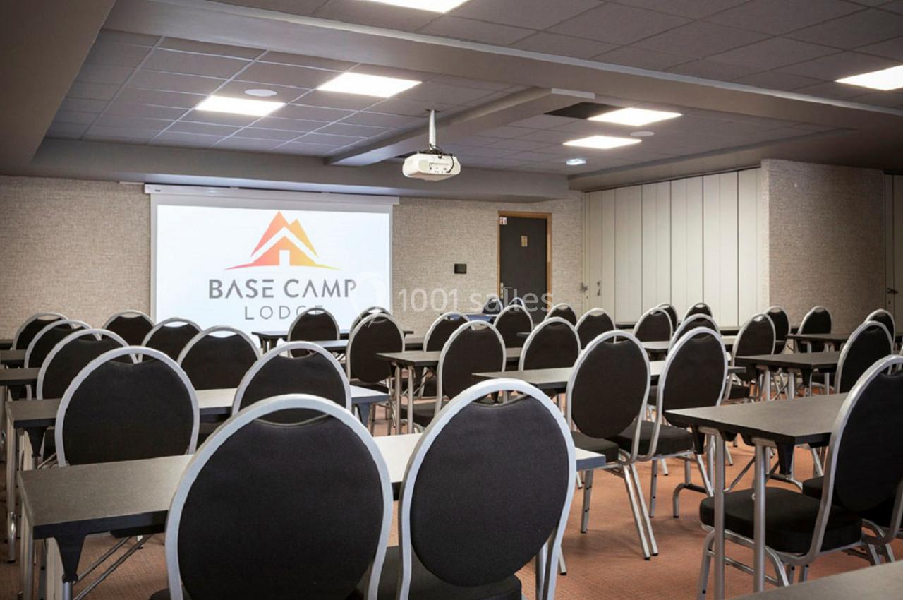 Salle de conférence avec des rangées de chaises noires et des tables, écran de projection affichant un logo.