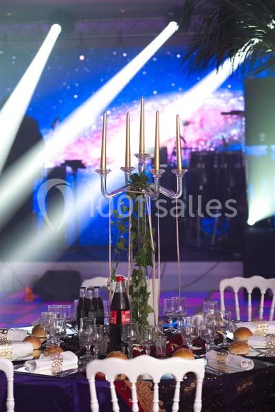 Location salle Le Raincy (Seine-Saint-Denis) - Le Chalet des Pins #10 Table de réception décorée avec un chandelier, des boissons, des assiettes et des pains, devant une scène éclairée.